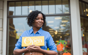 Black woman standing in front of her shop, holding a "open" sign